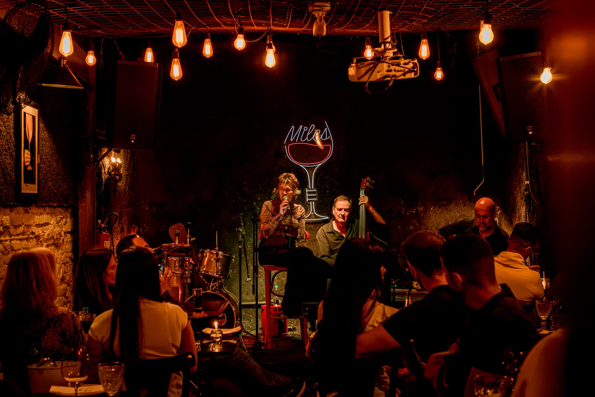 Salão com lâmpadas acesas ao som de jazz ao vivo, sob o letreiro neon Miles